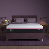 An Elite Series Mark Latex Foam Mattress by NapQueen, shown in a bedroom setting with a dark headboard and gray bedding, illuminated by bedside lamps.